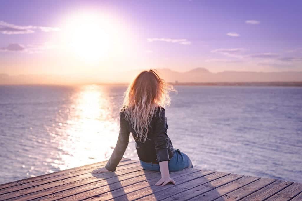 Back view of a woman who is sitting on a wooden pier with a beautiful view to the sunset. Sea, nature and sky in a one frame. Spain
